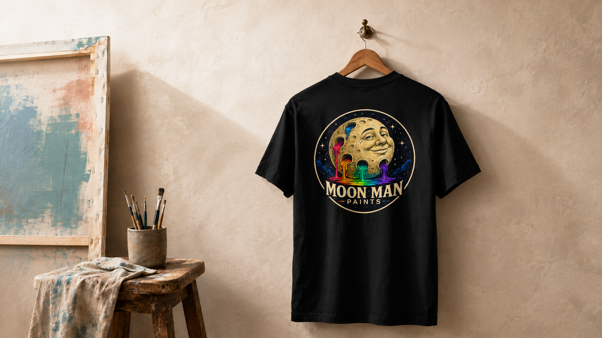 Black Moon Man Paints original logo tee displayed on an oak hanger in an artist studio